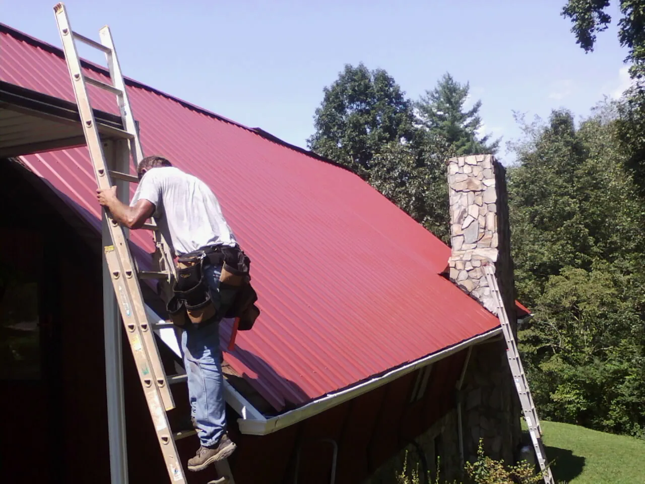 Roofer on a ladder beside a red metal roof and stone chimney — Soffit & Fascia Repair by Roofing Brothers in Warners